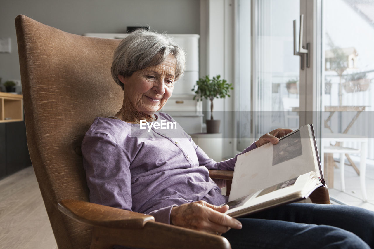 Portrait of senior woman sitting on armchair at home watching photo album