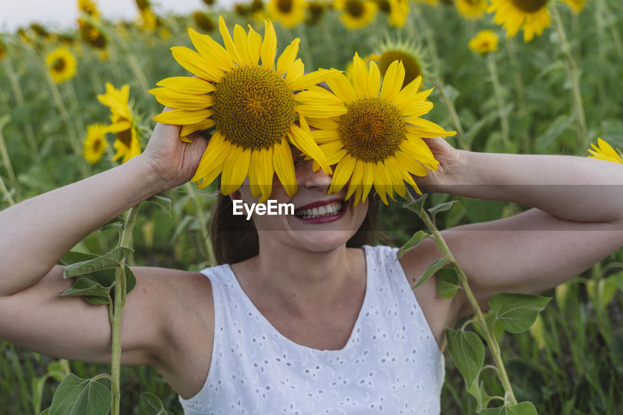 Happy woman covering eyes with sunflowers at field