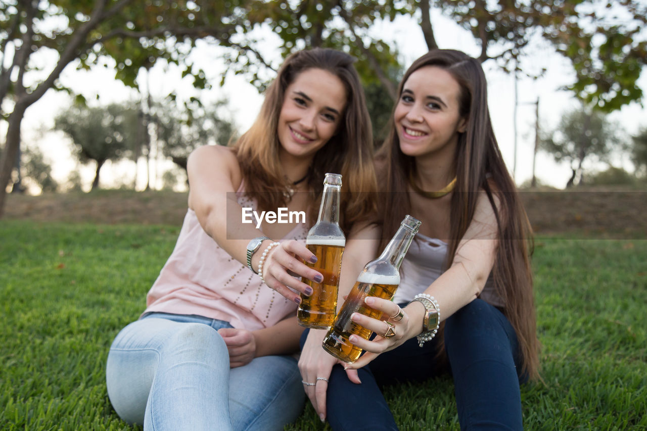 Smiling friends with alcoholic drinks in bottle sitting on grassy field at park