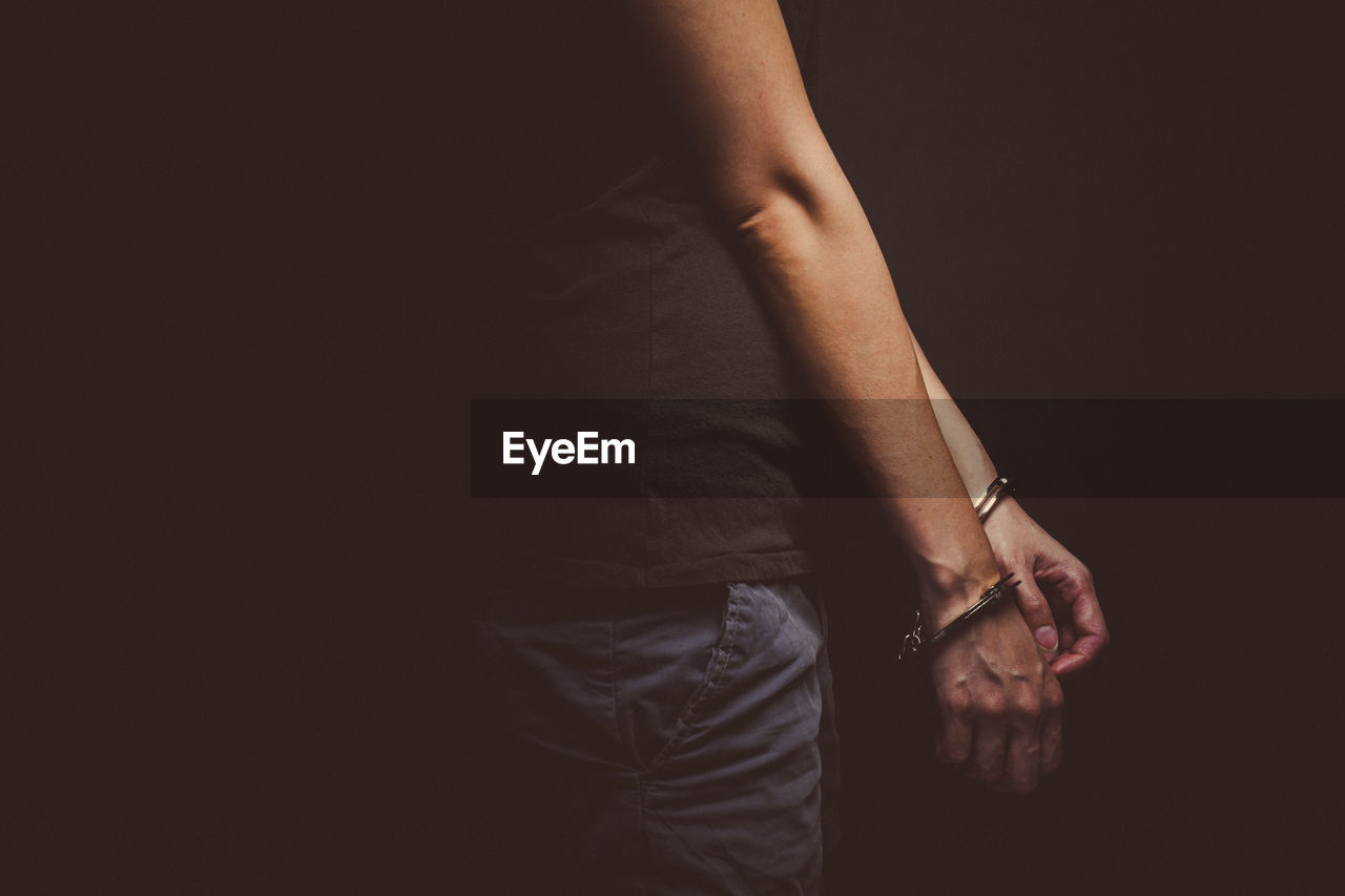 Midsection of man with handcuffs standing against black background