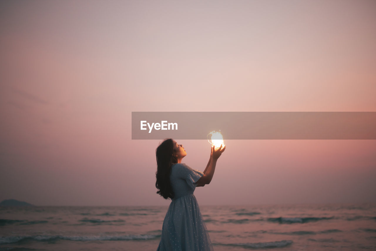 Side view of woman standing against sun during sunset