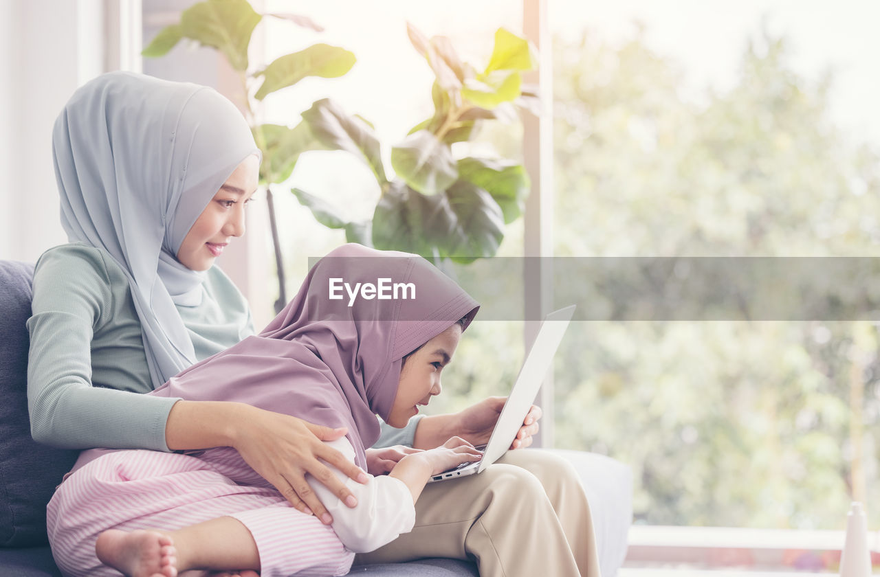 Asian muslim mother and daughter smiling when using laptop together, browsing internet or watching