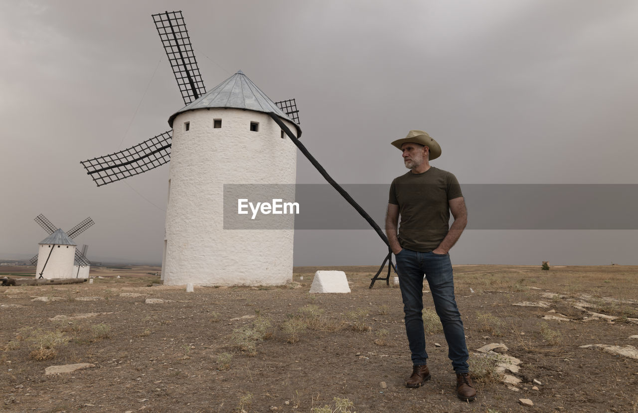 Man with cowboy hat standing by spanish traditional white windmills against sky