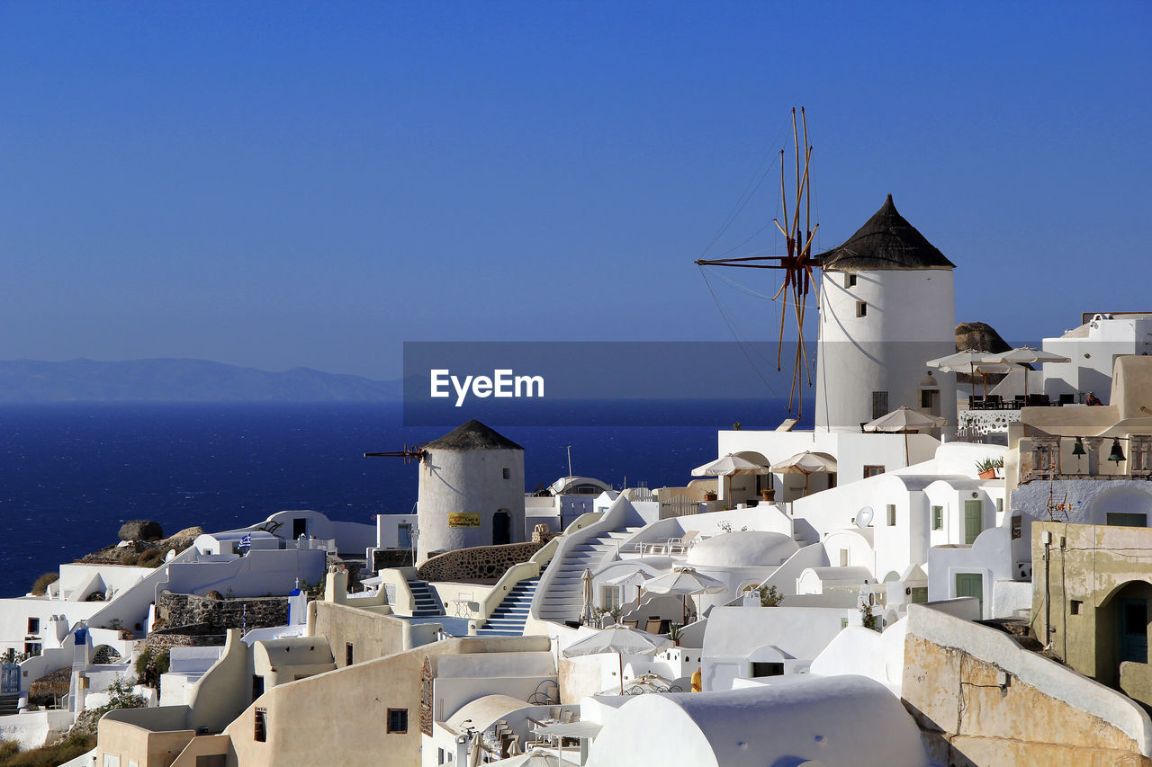 Traditional windmills amidst buildings on mountain by sea against clear blue sky