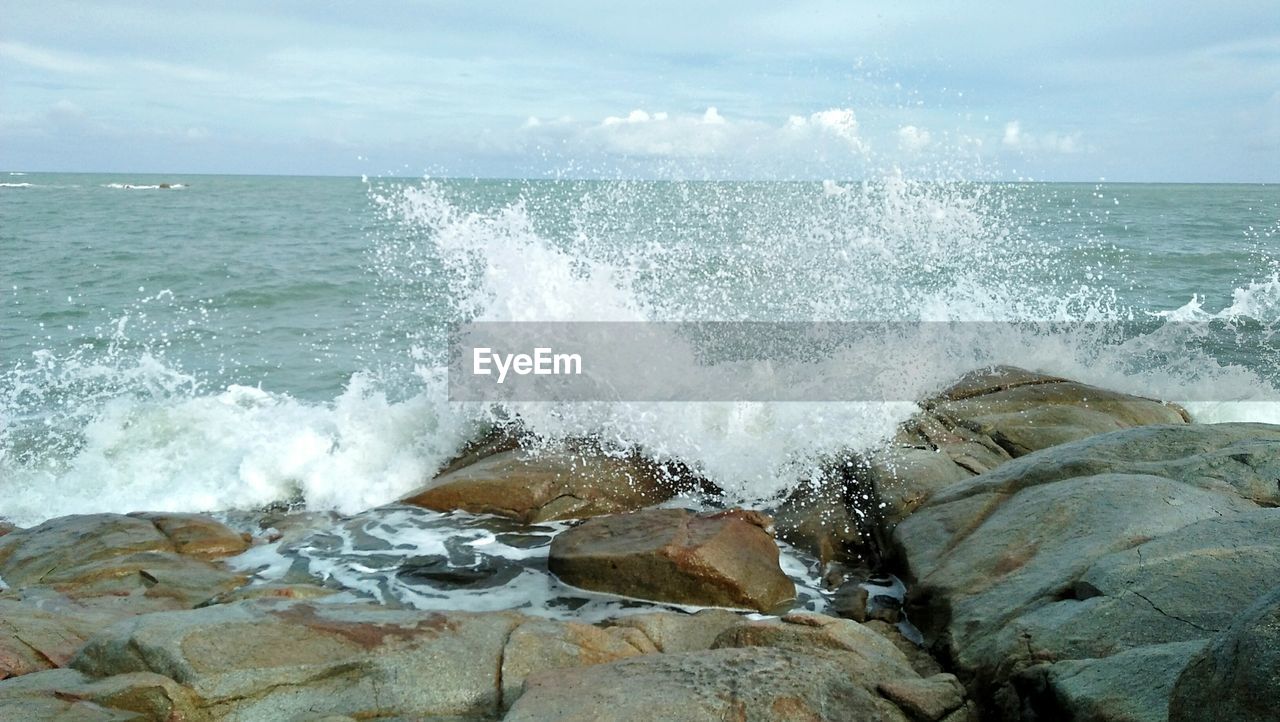 Waves splashing on rocks at shore against sky