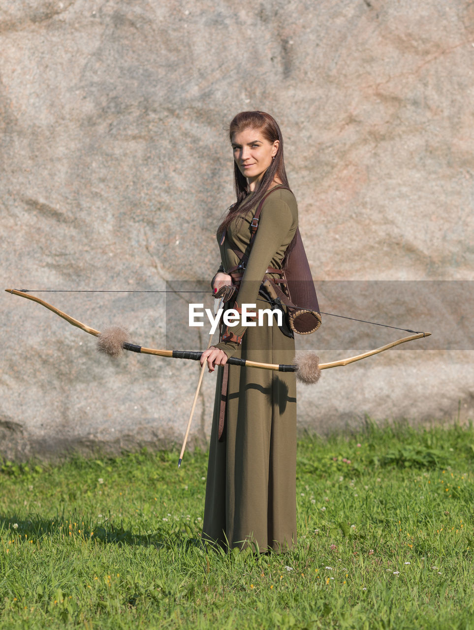 Portrait of woman holding bow and arrow standing on grass against rock