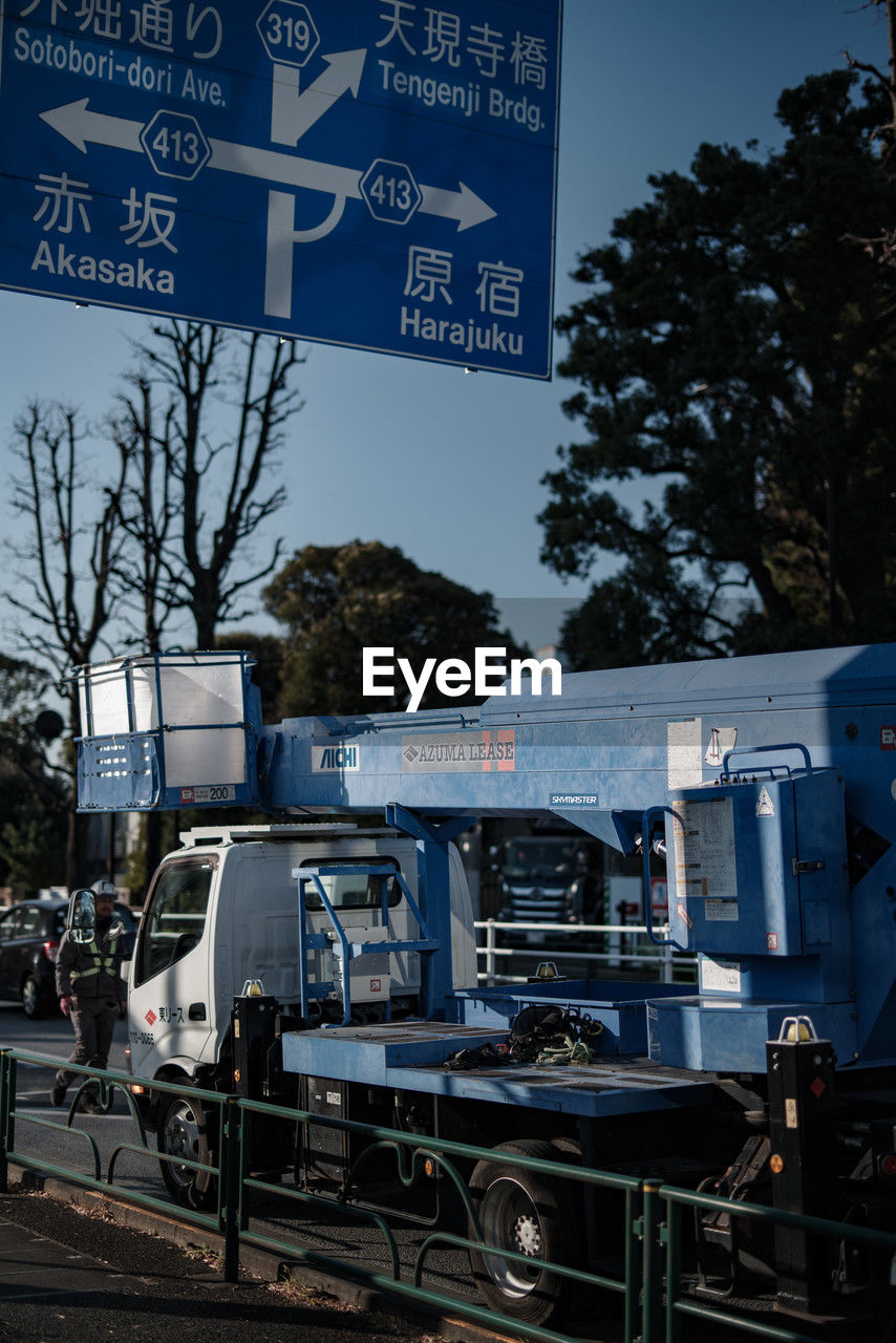 transportation, mode of transportation, tree, communication, sign, transport, text, nature, sky, vehicle, city, motor vehicle, plant, land vehicle, road, architecture, no people, outdoors, western script, car, street, day, urban area