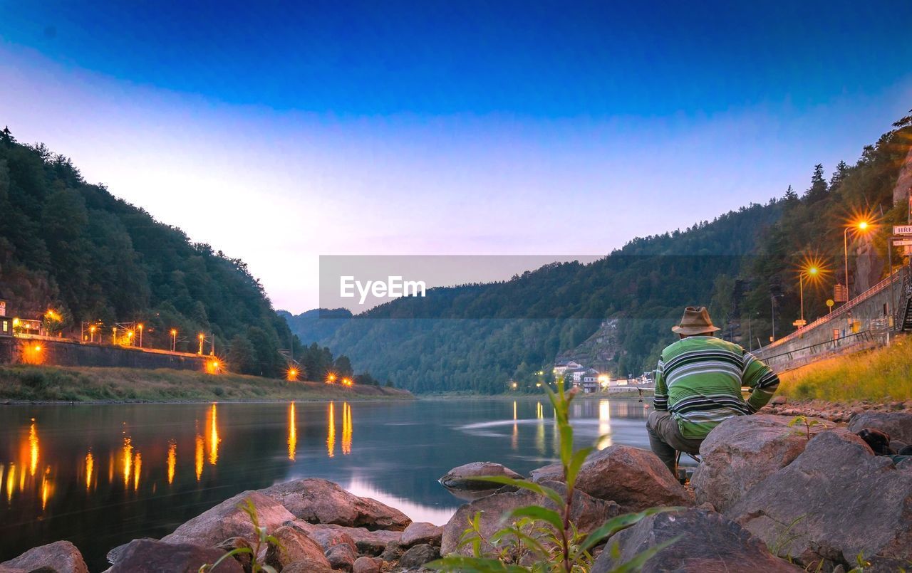 Rear view of man sitting by lake on rock