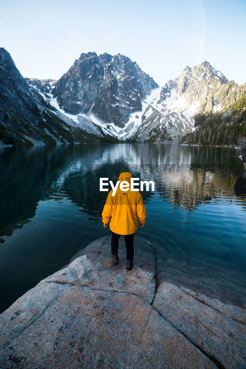 Rear view of man on rock by lake against mountains and sky