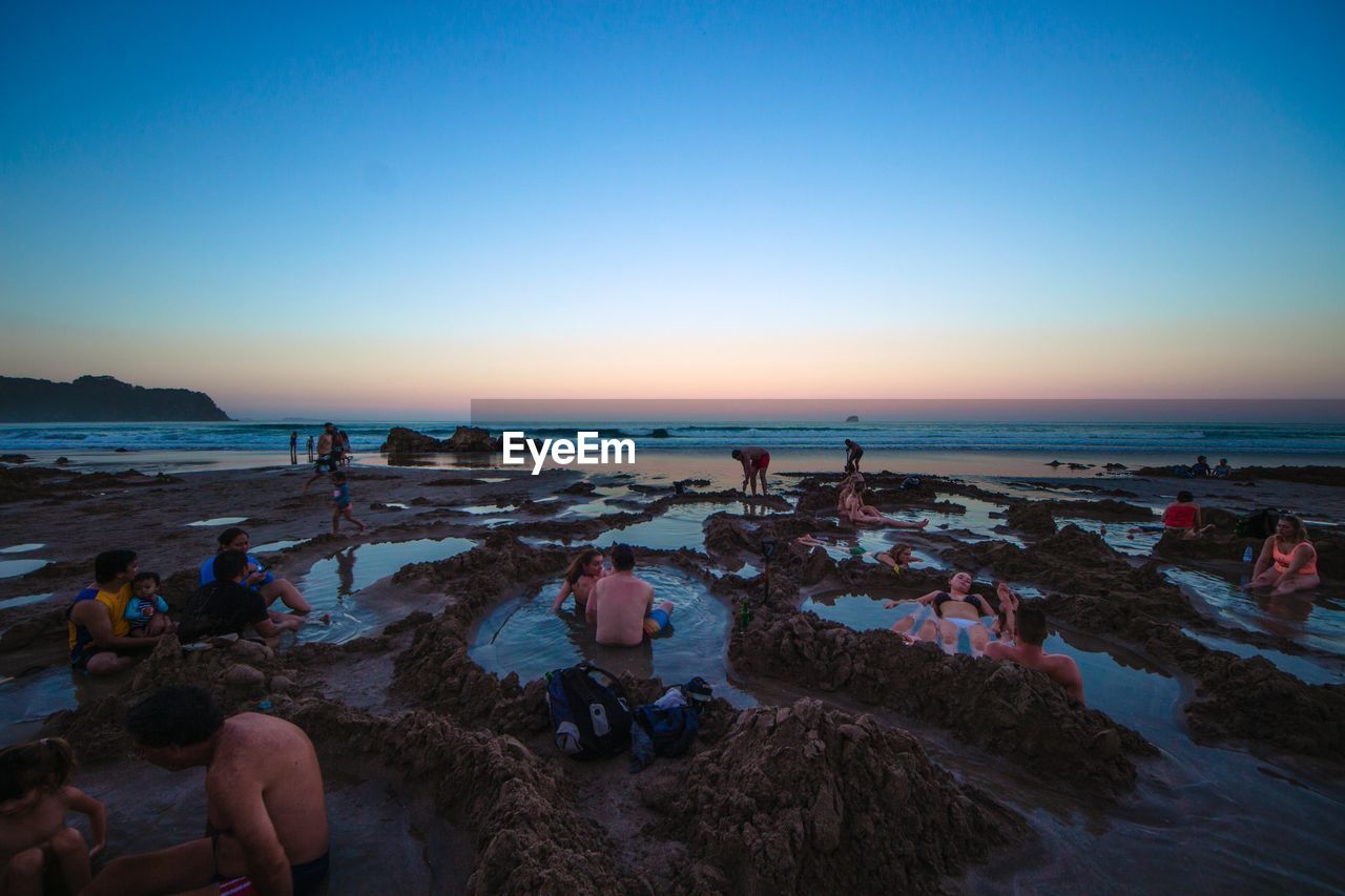 Couples enjoying at beach against clear blue sky during sunset