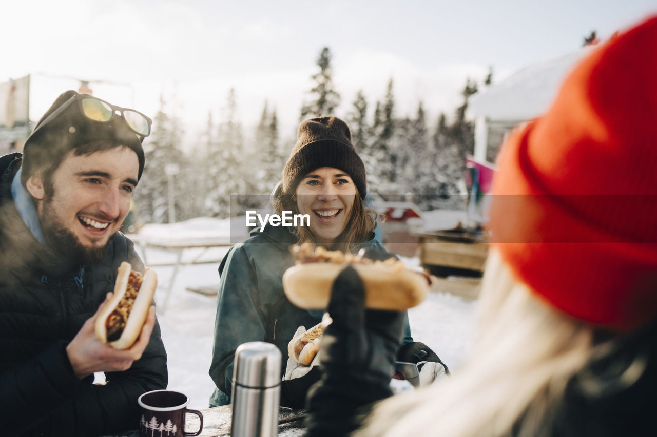 Happy friends eating hot dog while sitting at ski resort during winter