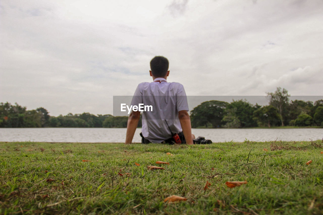 Rear view of man looking at lake while sitting on land against sky