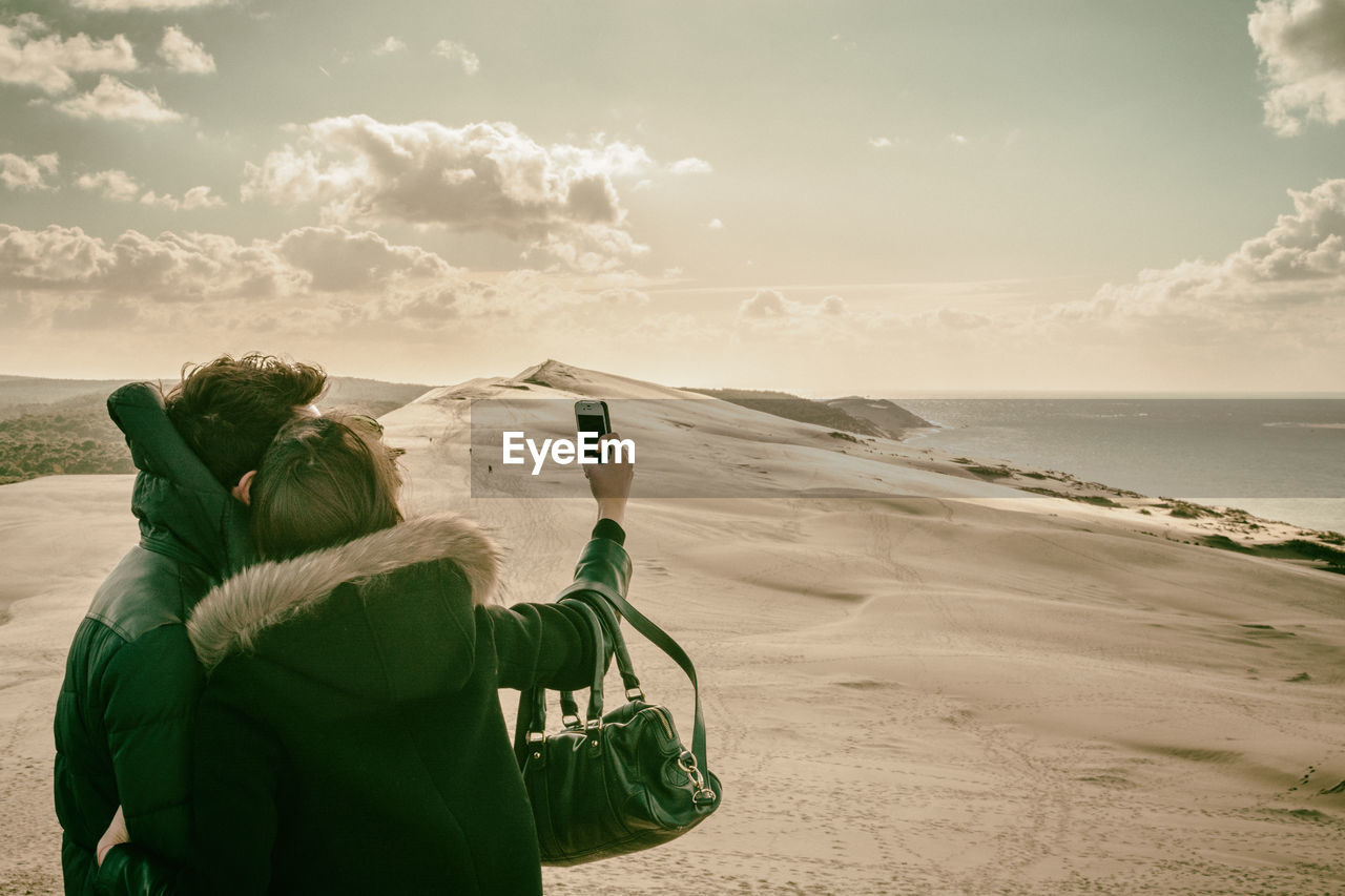 Couple taking selfie on sandy beach against sky