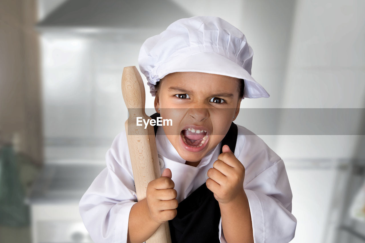 Portrait of girl wearing chef uniform screaming while holding rolling pin in kitchen