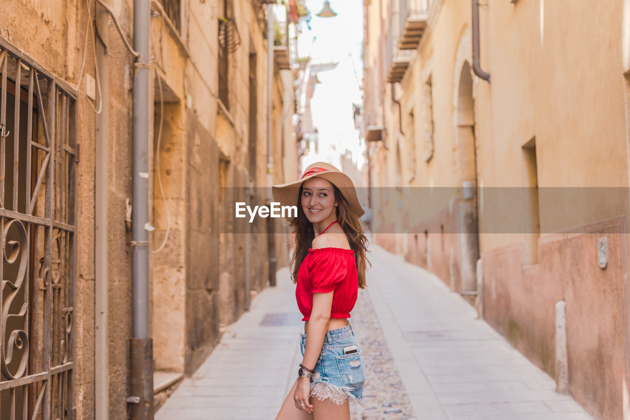 Smiling young woman wearing hat while standing in alley amidst buildings