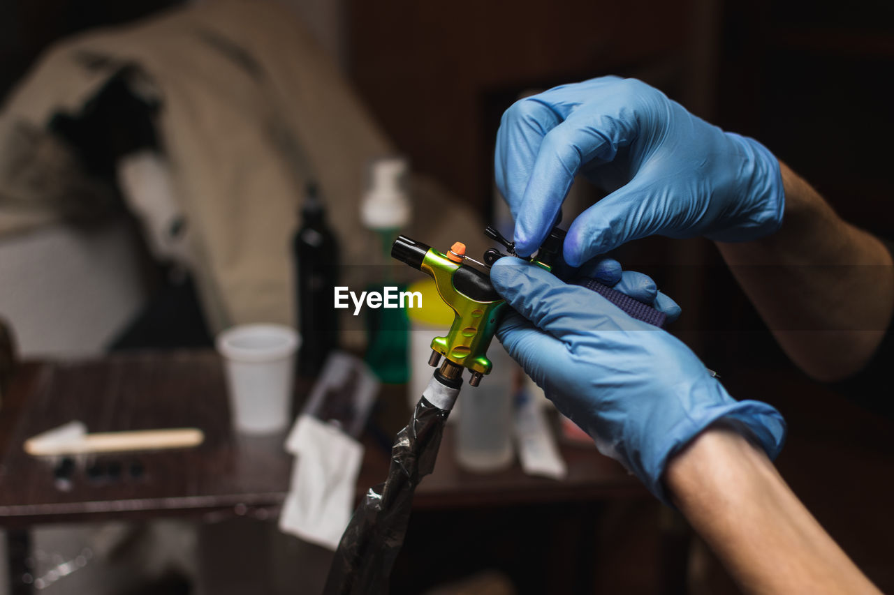 Side view of crop tattooist in protective gloves holding professional tattoo machine while working in salon