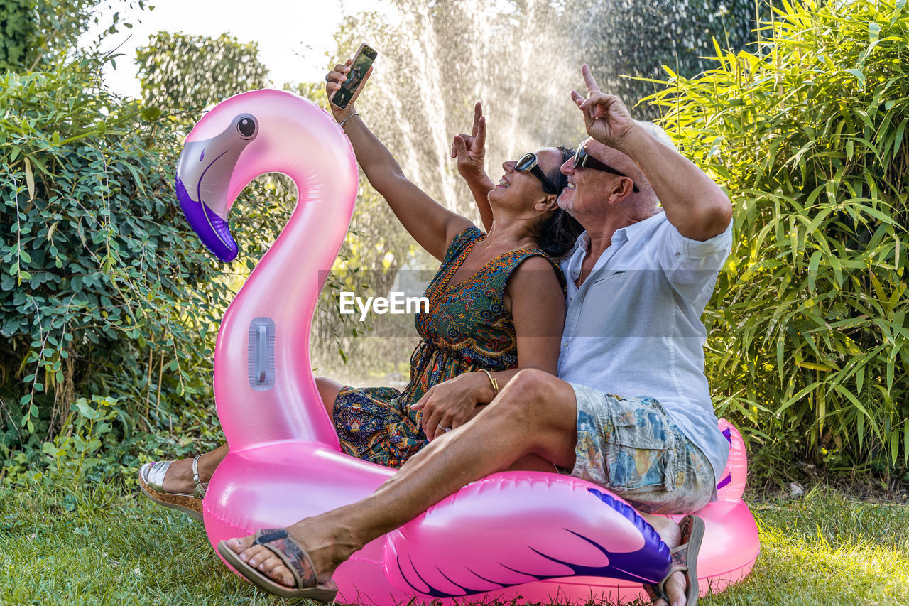 Beautiful smiling middle aged couple having fun taking a selfie sitting on flamingo inflatable toy