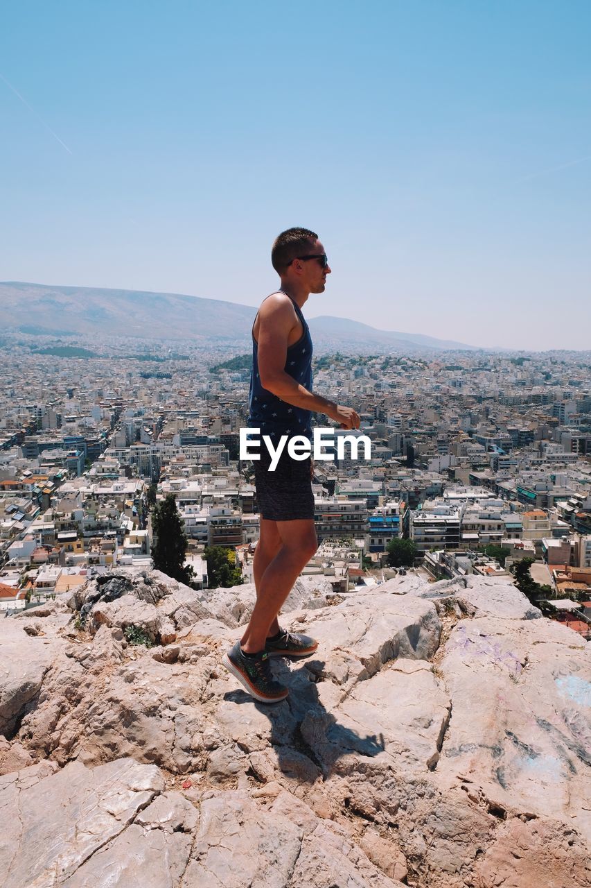 Side view of man overlooking cityscape while standing on mountain during sunny day