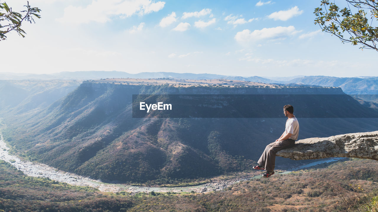 Man sitting on cliff while looking at mountains against sky