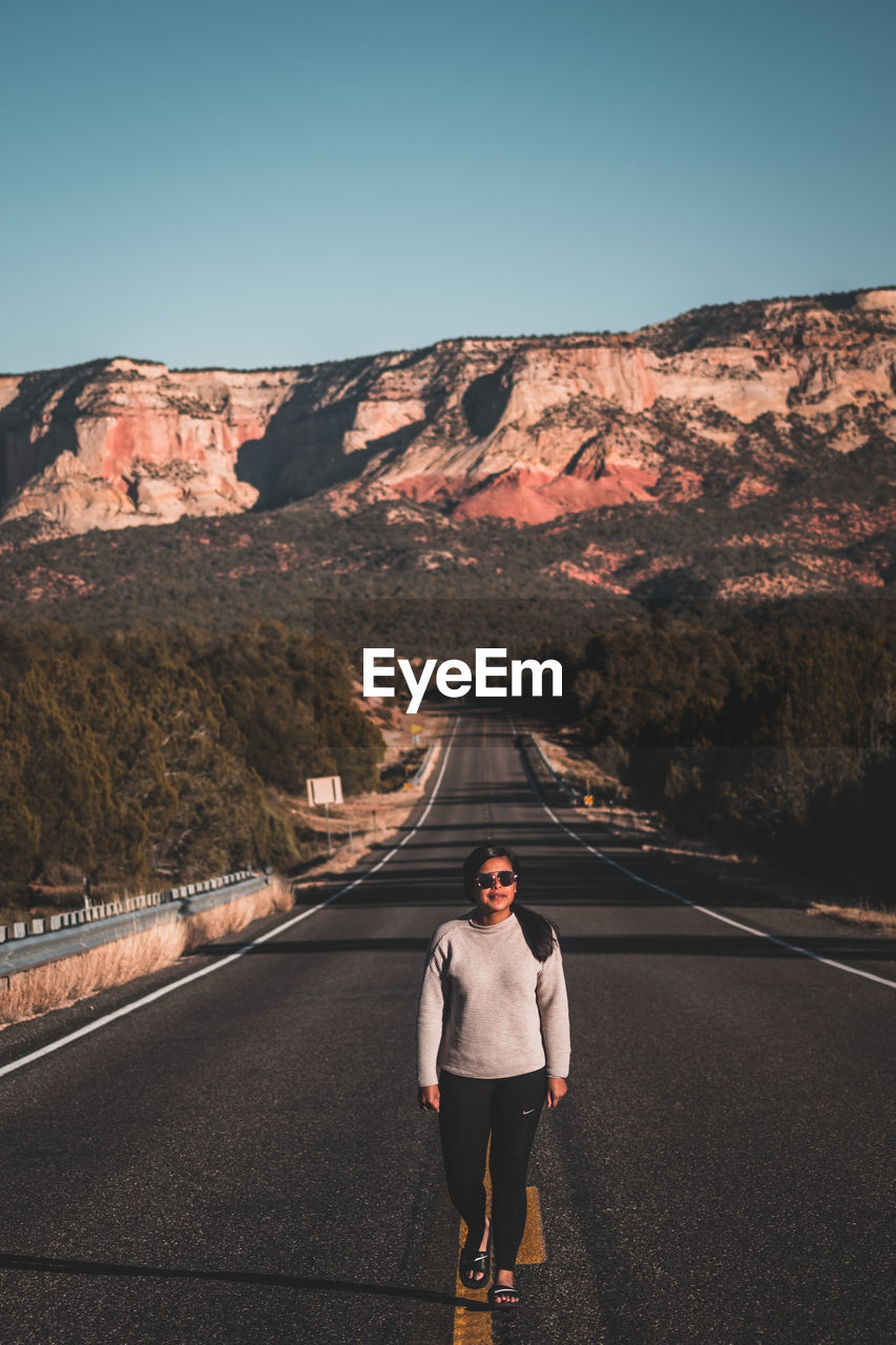 REAR VIEW OF WOMAN STANDING ON ROAD AGAINST MOUNTAIN