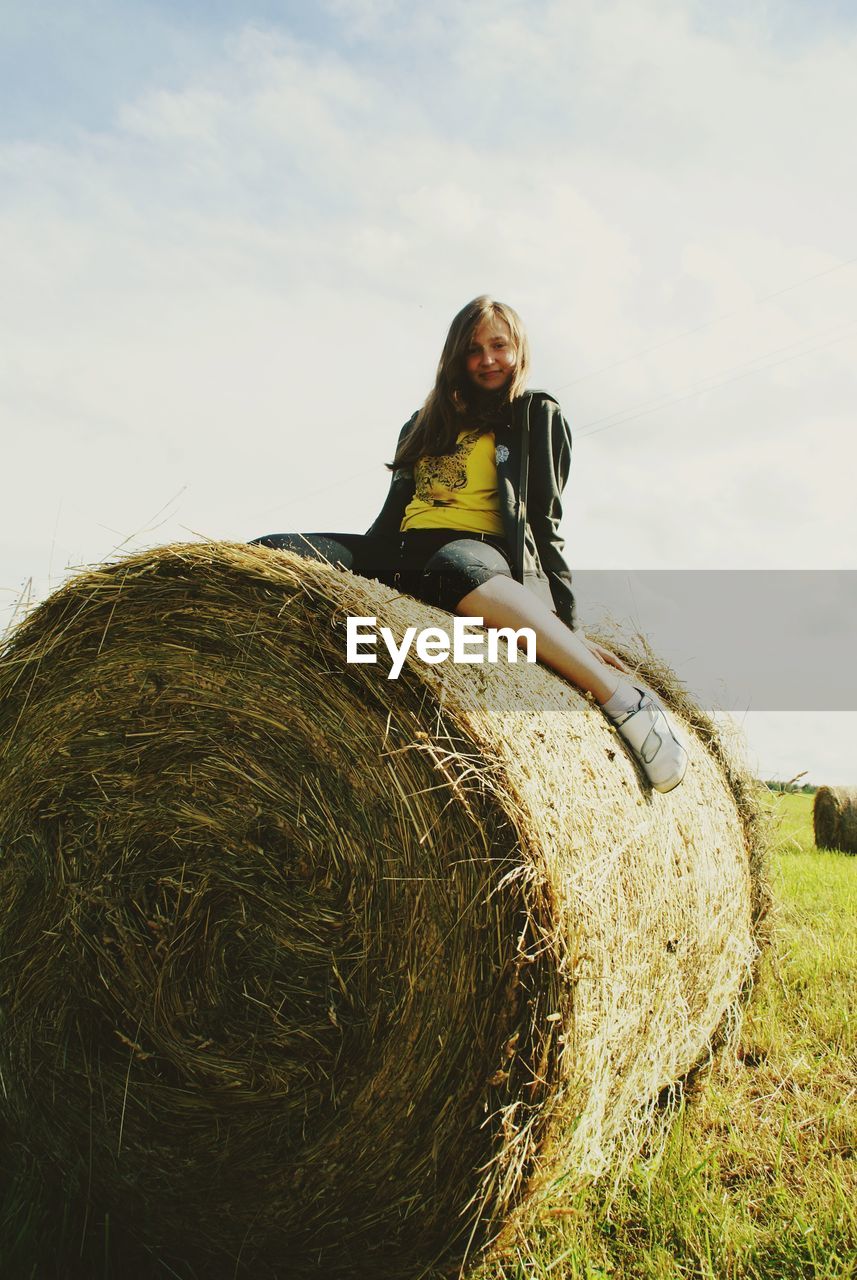 Young woman sitting on hay bale at field against sky during sunny day