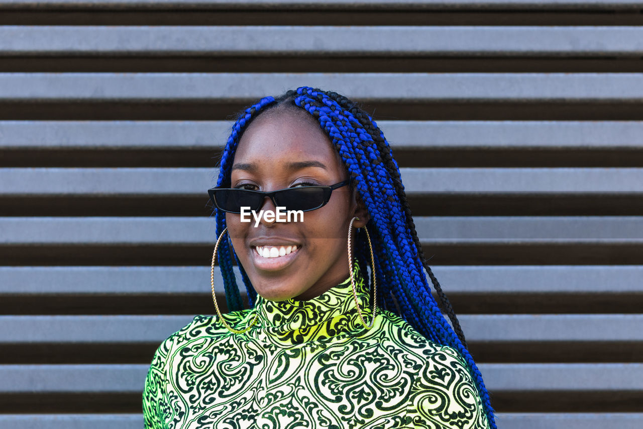 Confident happy african american female with blue braids wearing trendy outfit and sunglasses standing on street and looking at camera