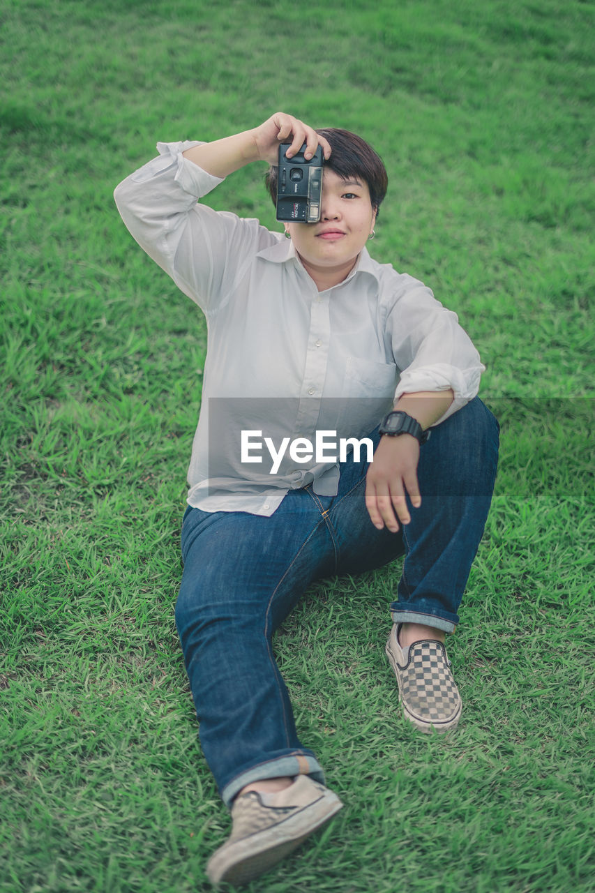 Portrait of young woman with camera sitting on grassy land in park