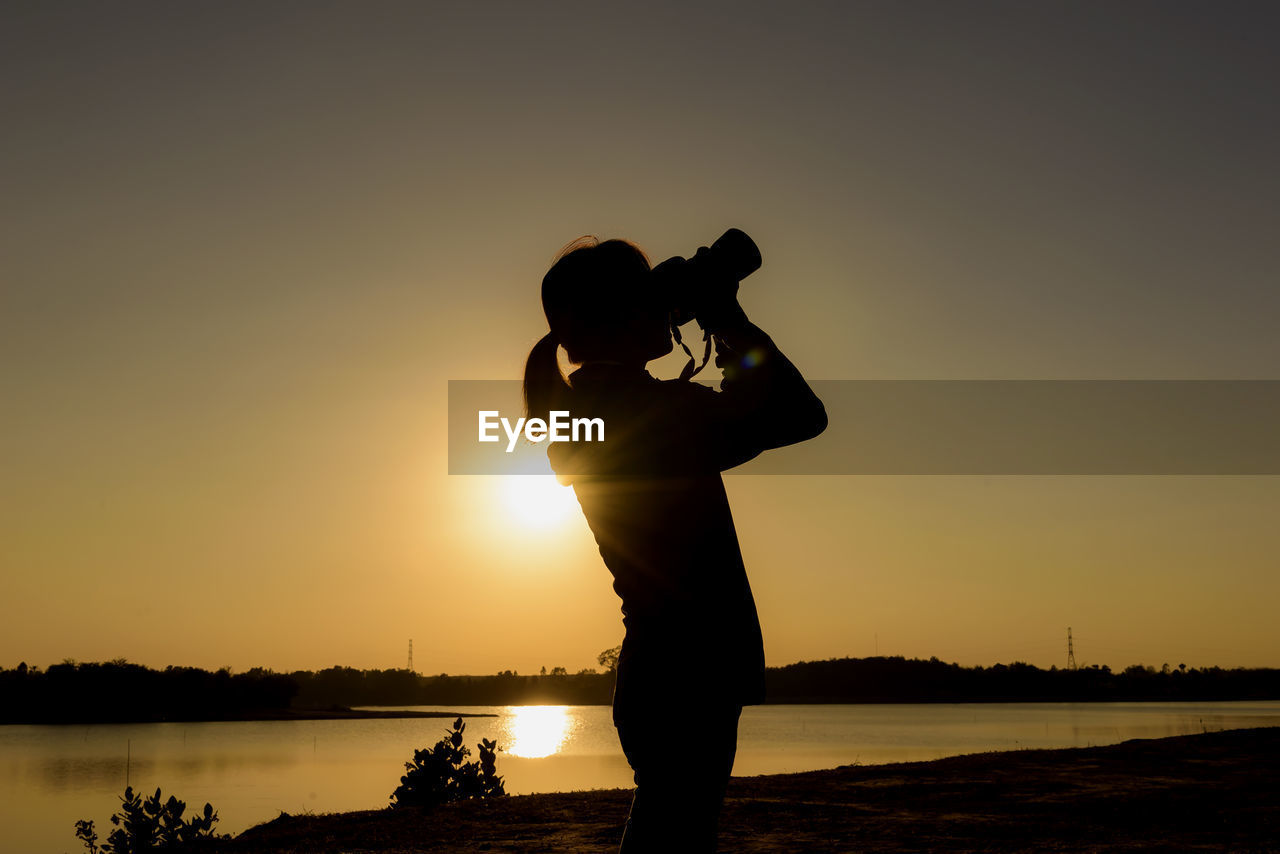 Silhouette man photographing lake against sky during sunset