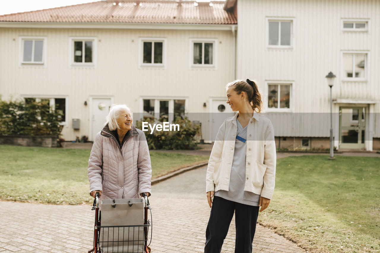 Smiling female nurse and senior woman looking at each other while walking against house