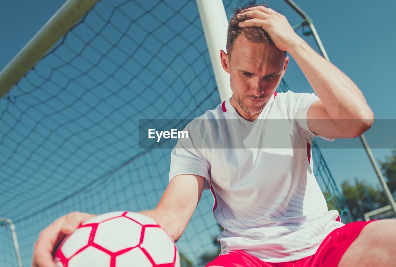 Low angle view of worried soccer player sitting by goal post against sky