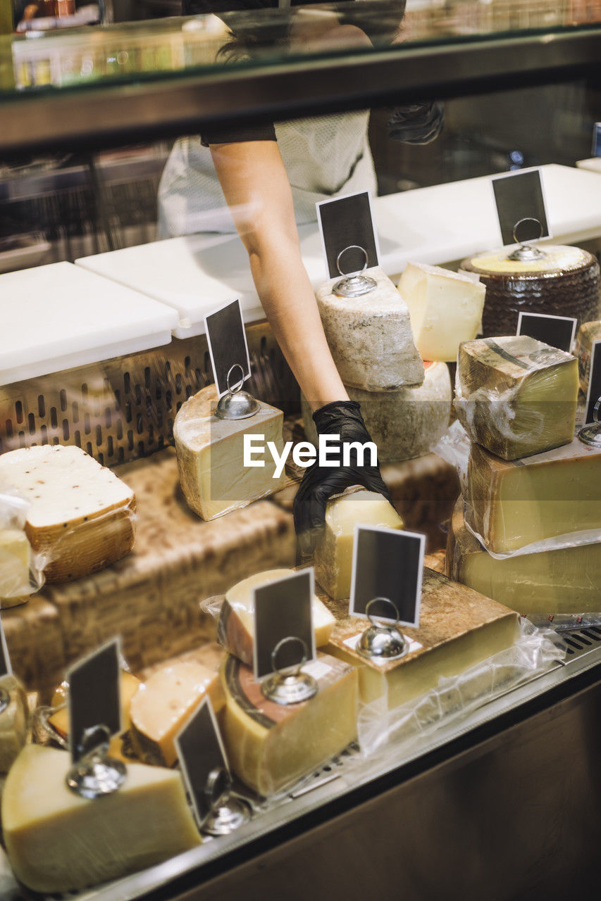 Midsection of female retail clerk removing cheese from display cabinet at grocery store