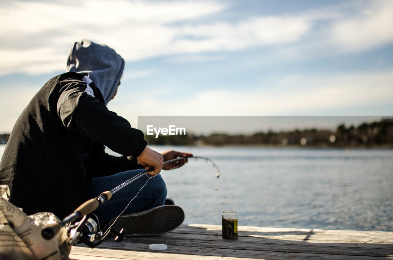 Man fishing while sitting on pier over lake against sky