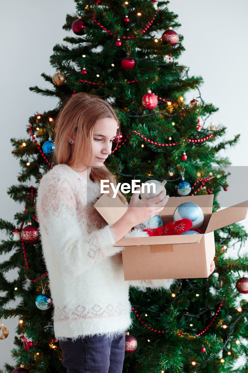 Teenage girl looking at christmas ornament while sitting by christmas tree at home