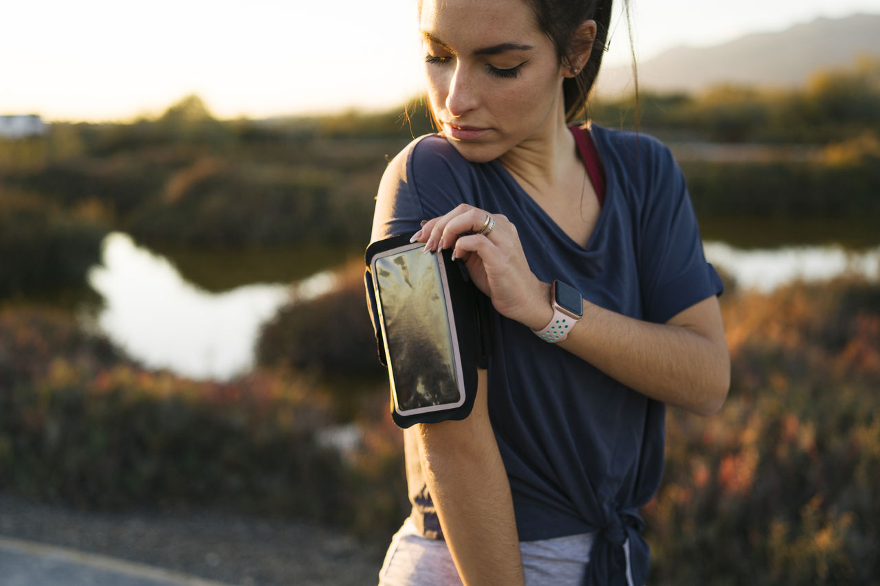 Young woman adjusting smart phone in arm band while standing against landscape