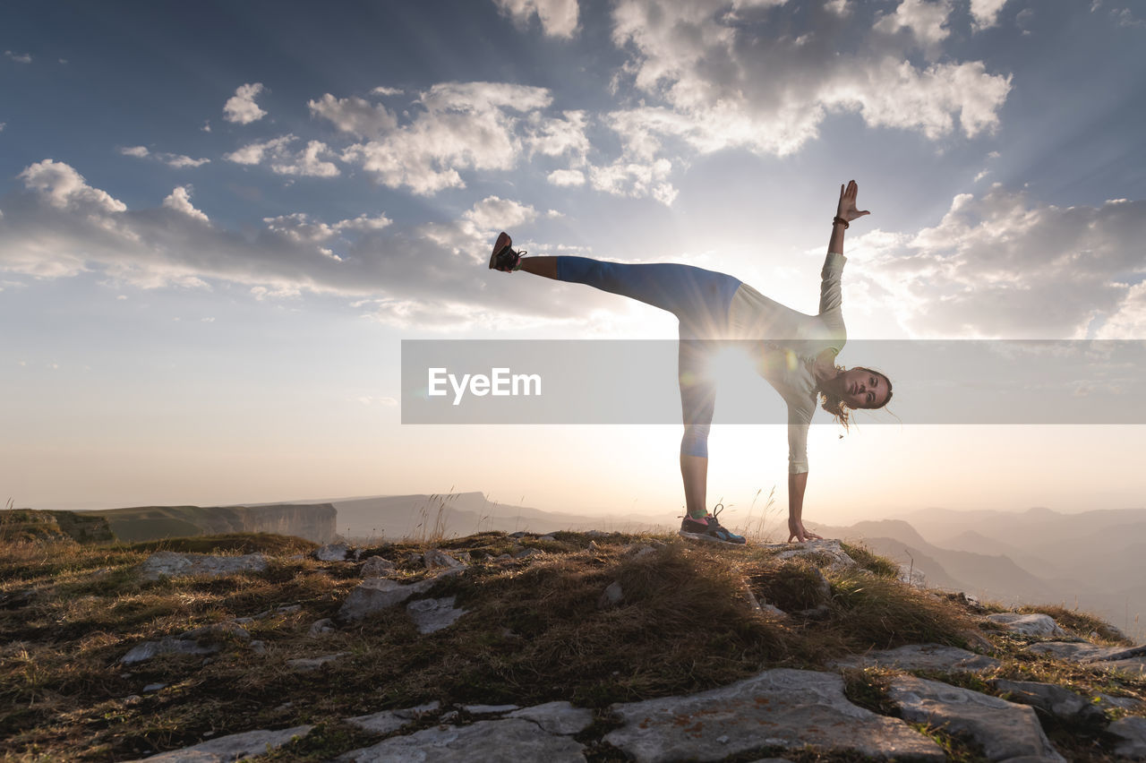 Young woman doing beautiful yoga, sunset or sunrise in mountains over blue sky and clouds against