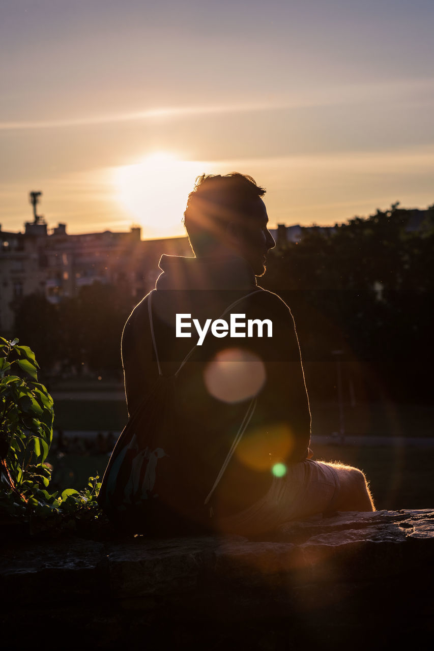 Man sitting in berlin on wall against sky during sunset with lens flare and a park in the background