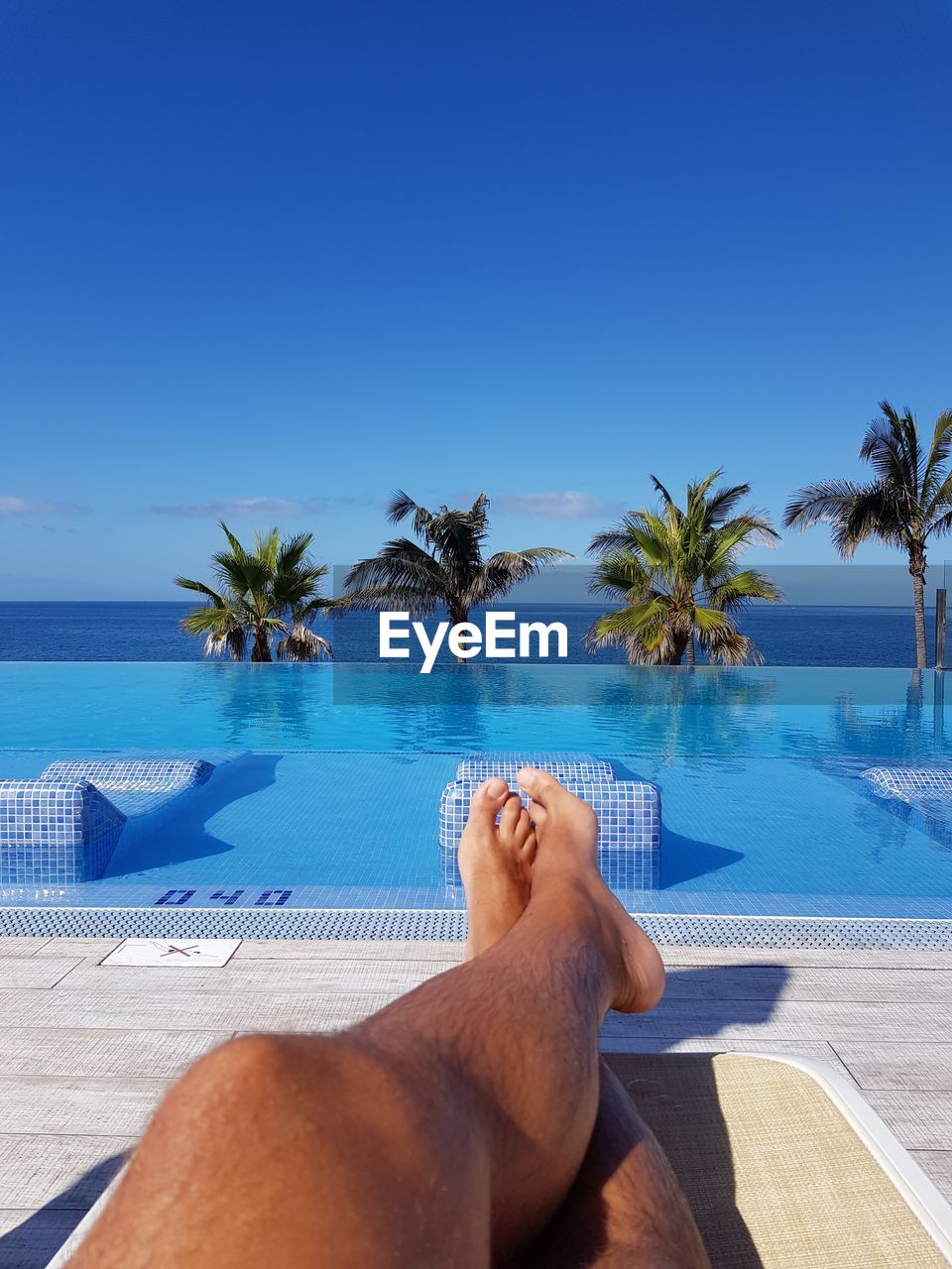 Low section of man relaxing on deck chair by infinity pool against blue sky