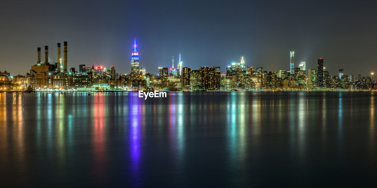 Illuminated cityscape by sea with reflection against sky during night