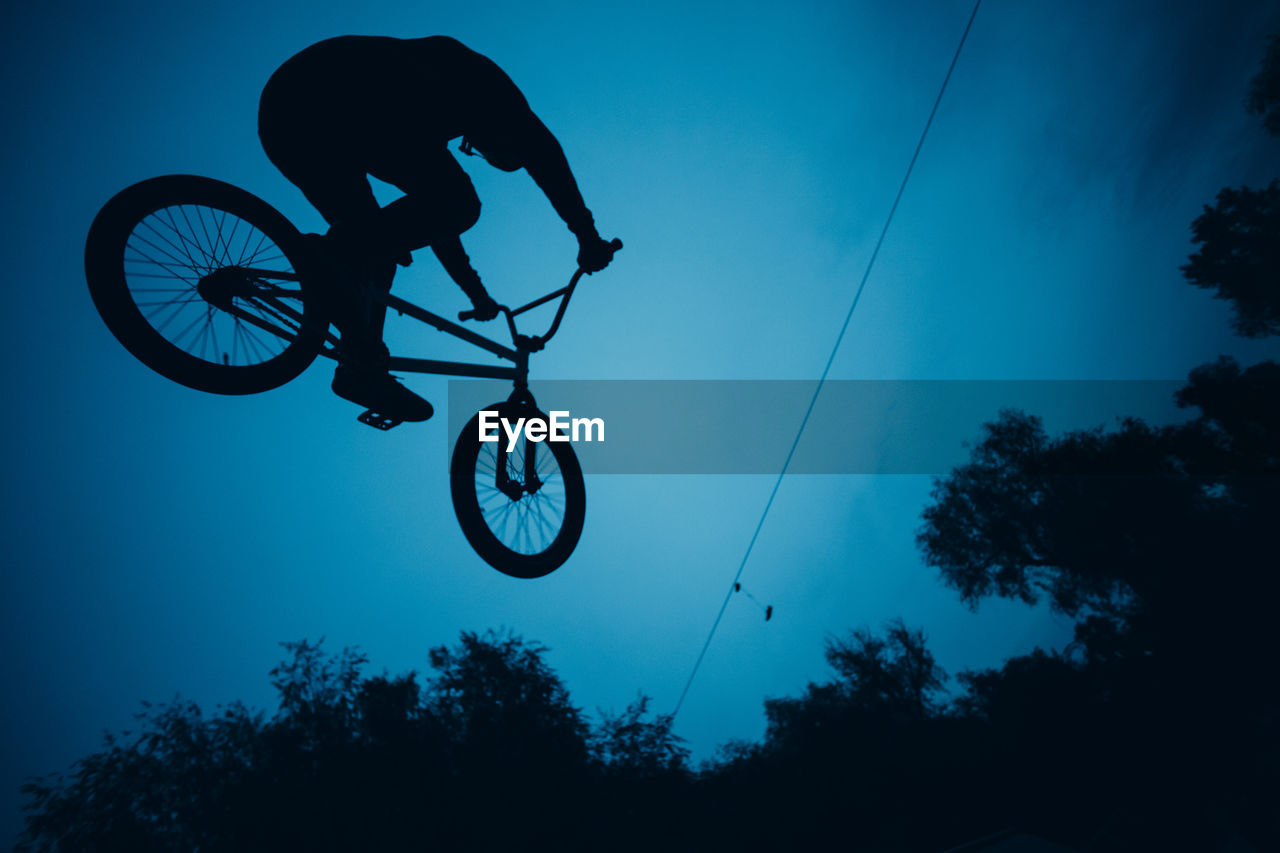 Low angle view of silhouette person flying bicycle against sky