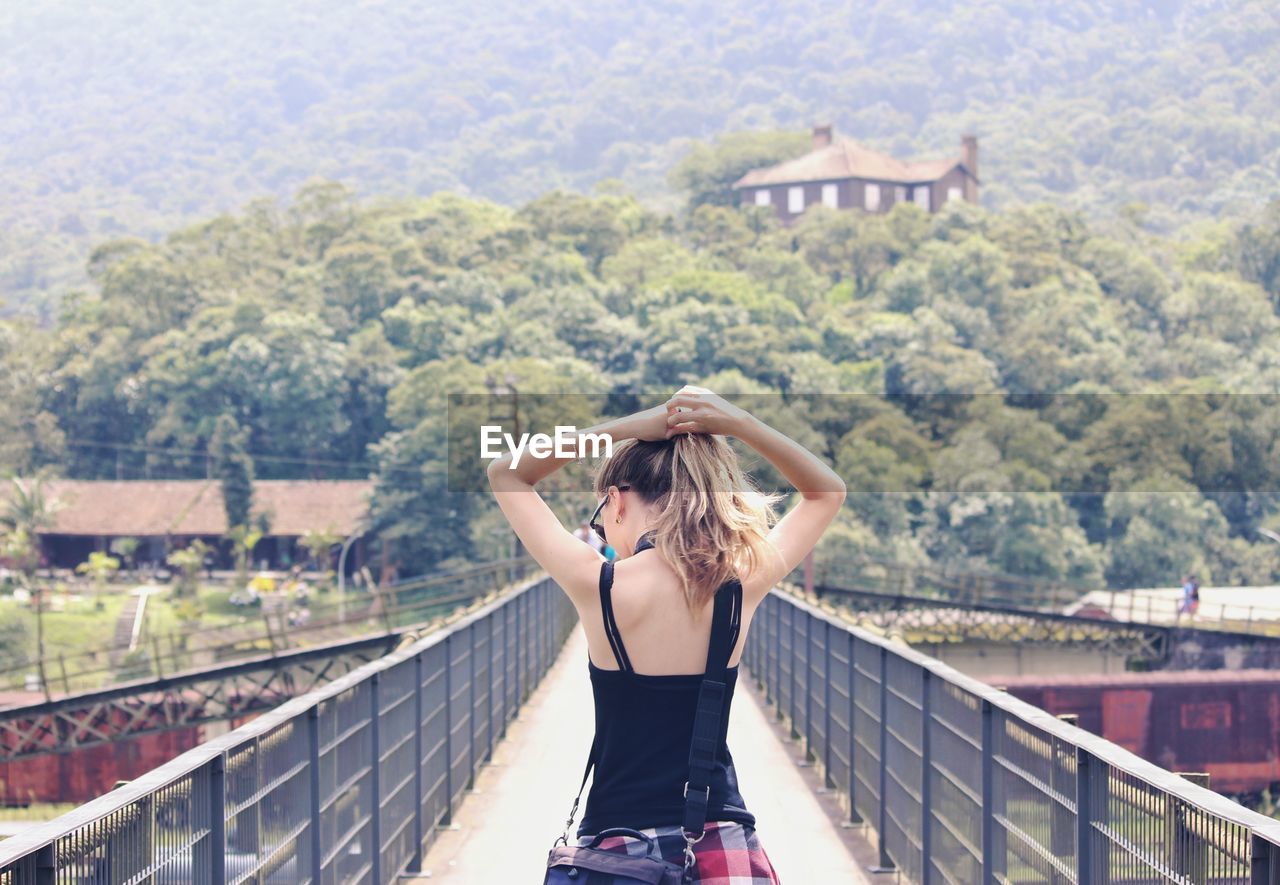 Woman tying hair while walking on footbridge against trees