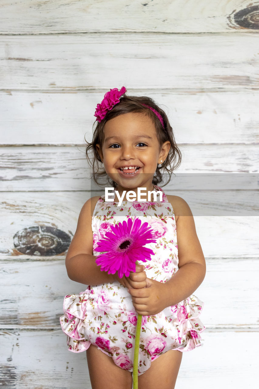 Portrait of smiling girl holding pink flower against wooden wall
