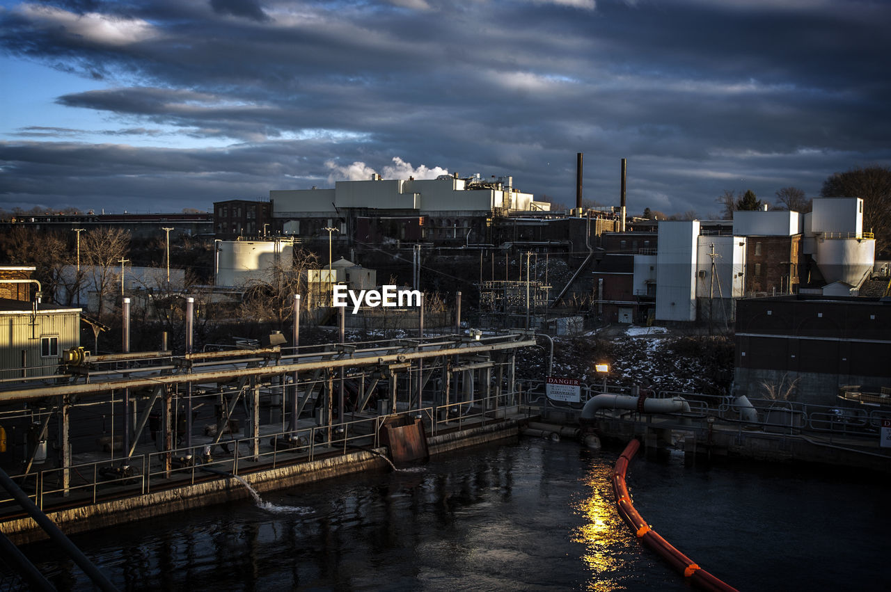 Factory by river against cloudy sky at dusk