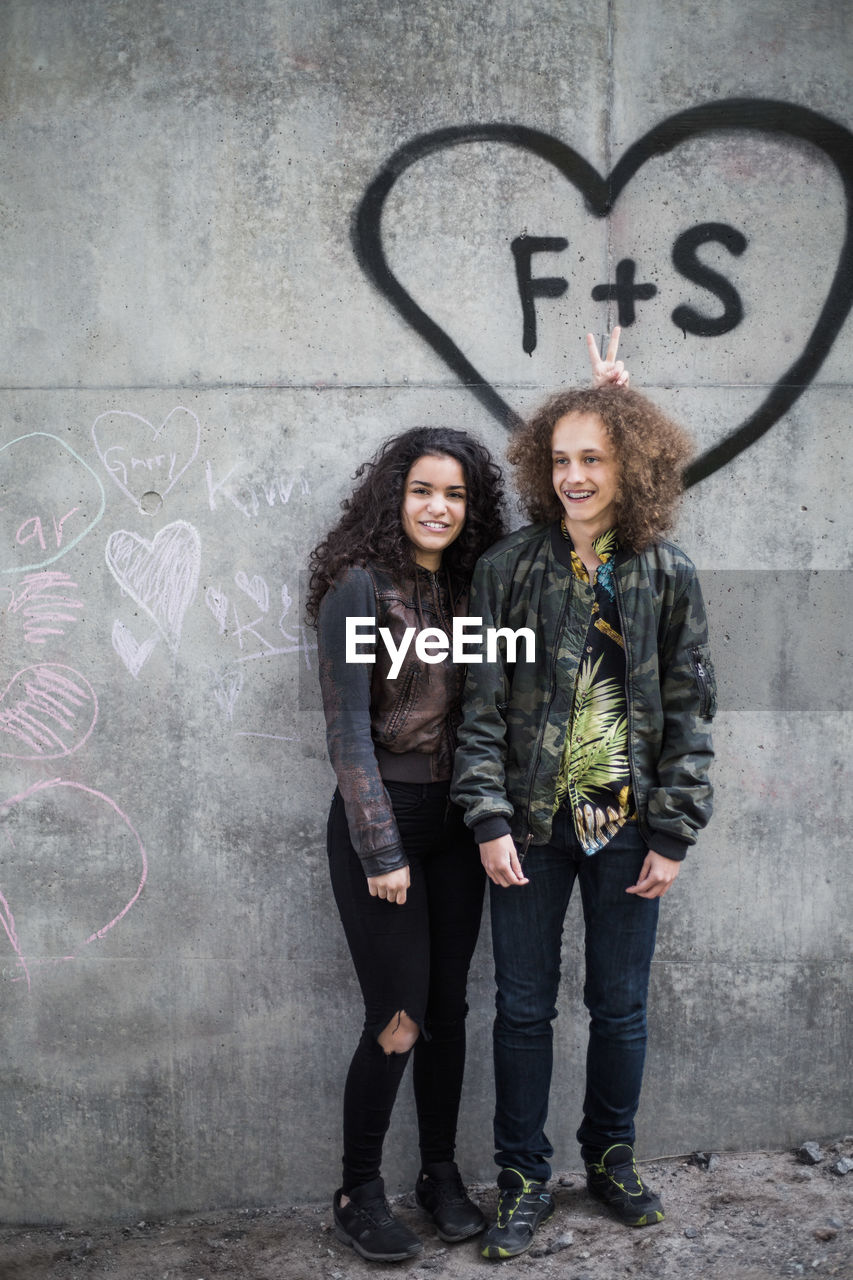 Cheerful teenage friends standing against graffiti on gray wall in city