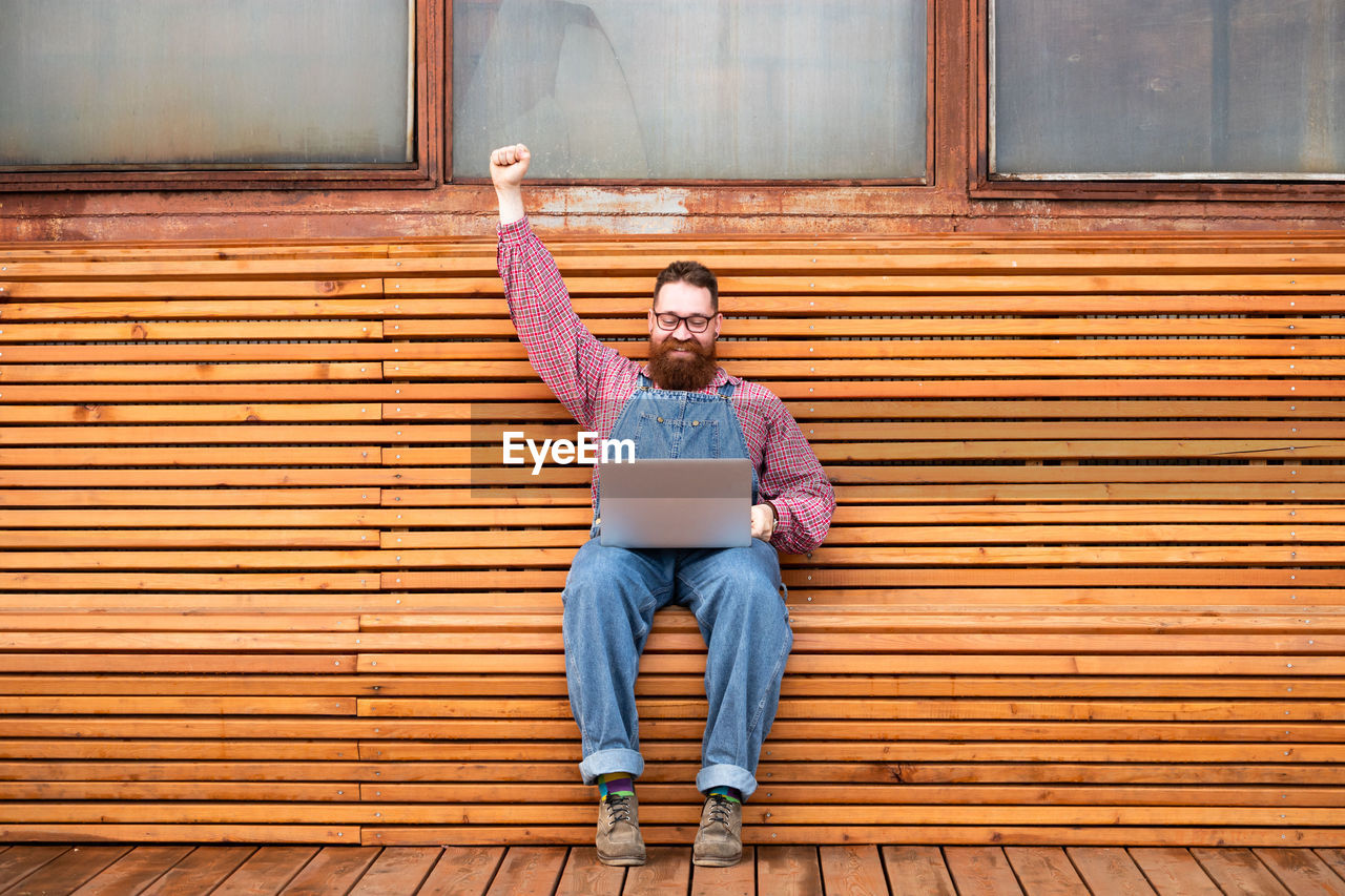 Cheerful brutal bearded hipster man working on laptop sitting on bench rising hand up celebrating.