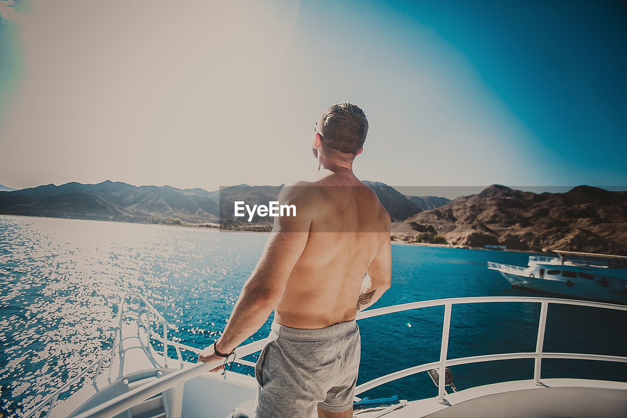 Shirtless man standing on boat in sea against sky