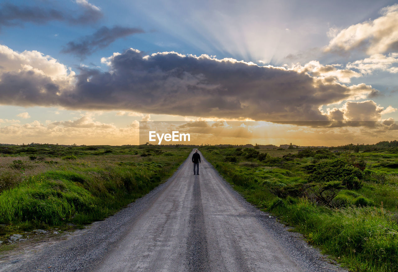 Man standing on road amidst landscape against sky during sunset