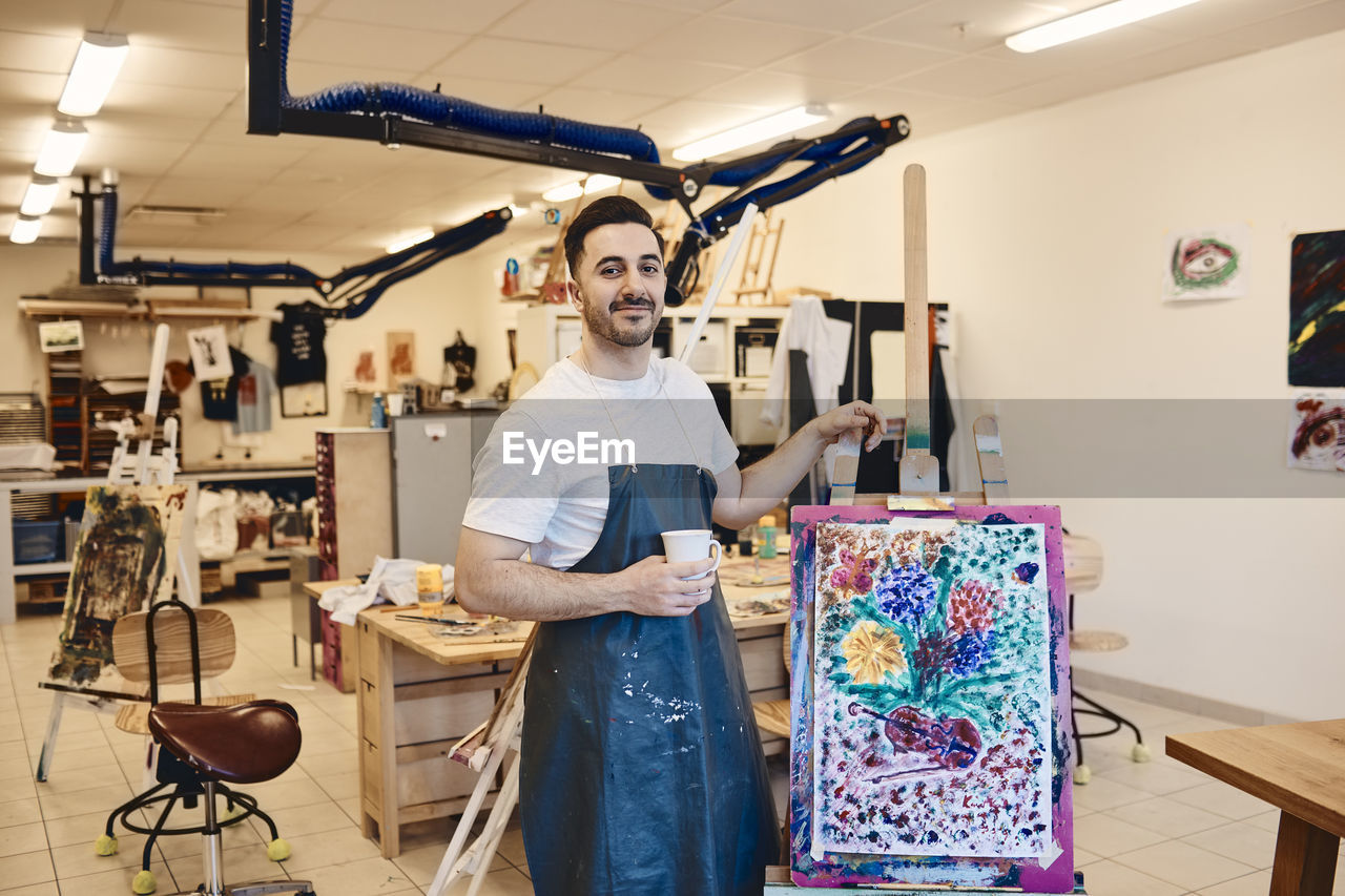 Portrait of confident male artist standing by easel with painting in art class