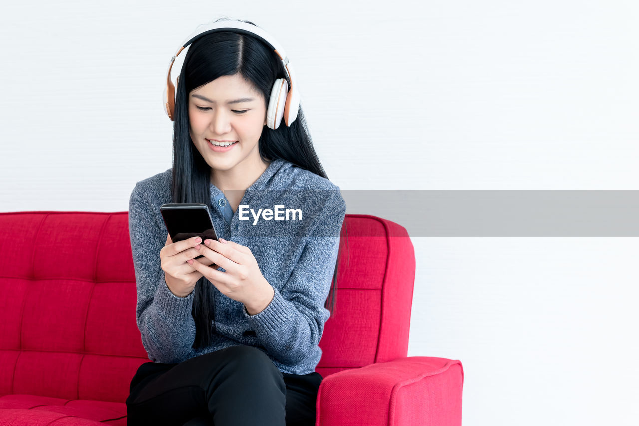 SMILING YOUNG WOMAN USING PHONE WHILE SITTING ON WALL