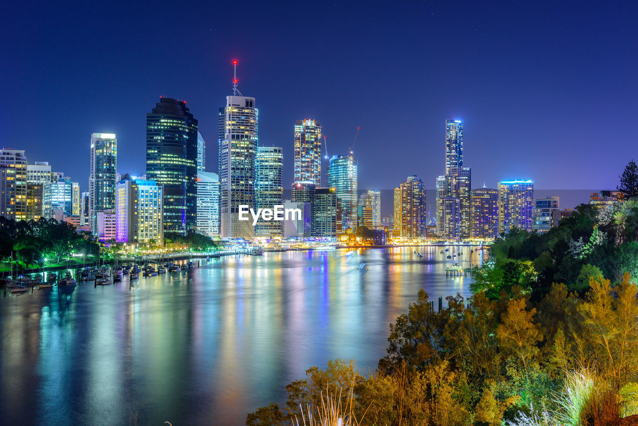 Illuminated buildings by river in city against sky at night