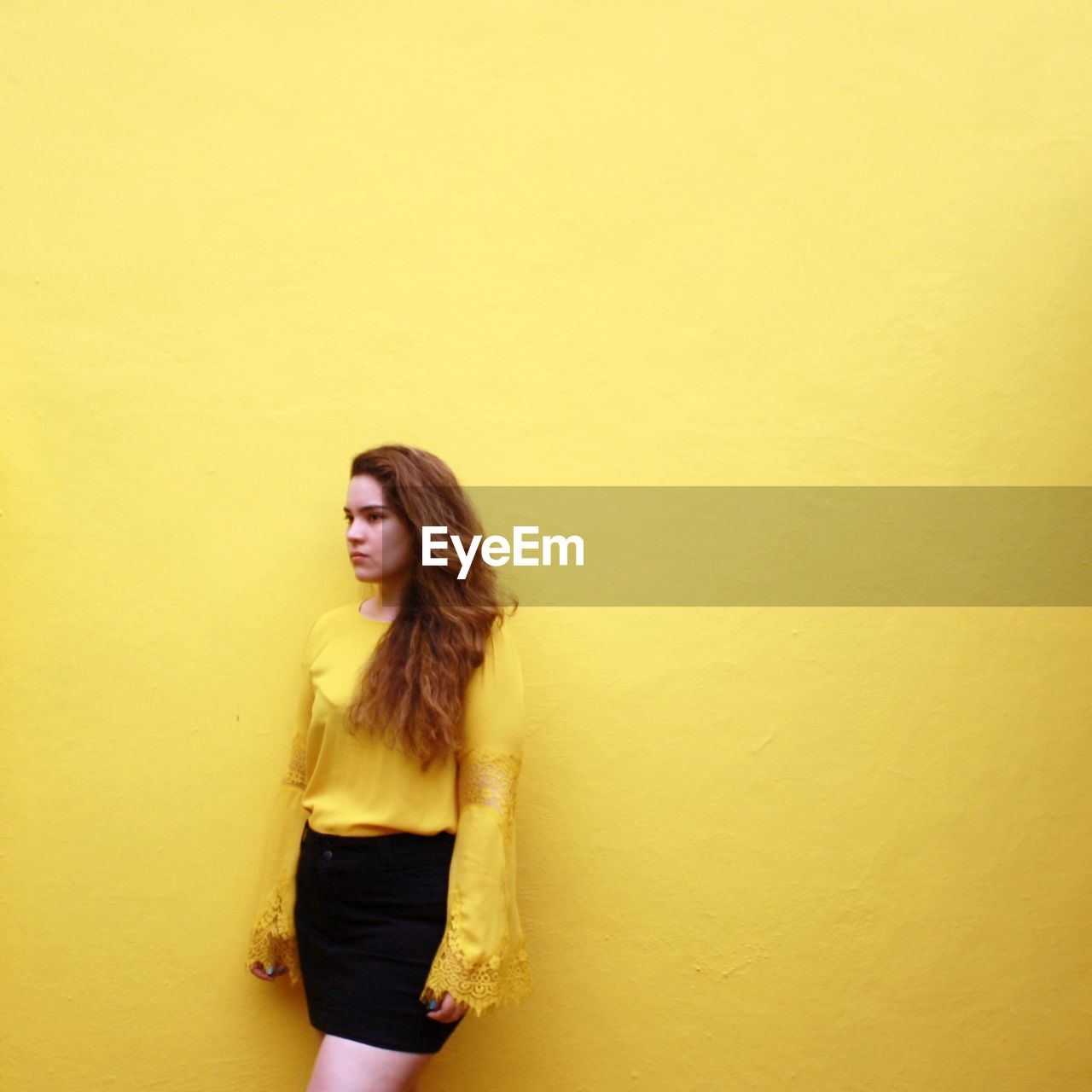 Young woman looking away while standing against yellow wall